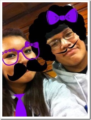 Photo of a boy and girl. Girl has purple glasses and a black moustache drawn on. The guy has a afro with a purple bow and a pencil moustache drawn on.